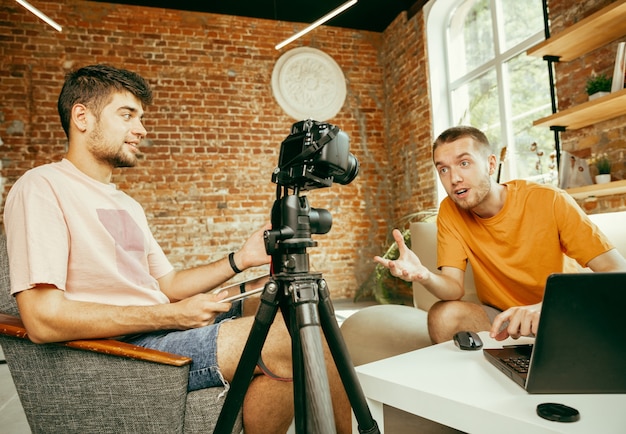 Dwóch Młodych Blogerów Rasy Kaukaskiej W Zwykłych Ubraniach Z Profesjonalnym Sprzętem Lub Kamerą Nagrywającą Wywiad Wideo W Domu. Blogowanie, Videoblog, Vlogowanie. Rozmowa Podczas Transmisji Na żywo W Pomieszczeniu.