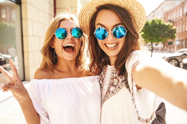 Dwóch Młodych Kobiet Uśmiechniętych Hipisów Brunetka I Blond Modelki W Letnim Białym Stroju Hipster Biorąc Selfie Zdjęcia Do Mediów Społecznościowych Na Telefon. Zaskocz Twarz, Emocje,