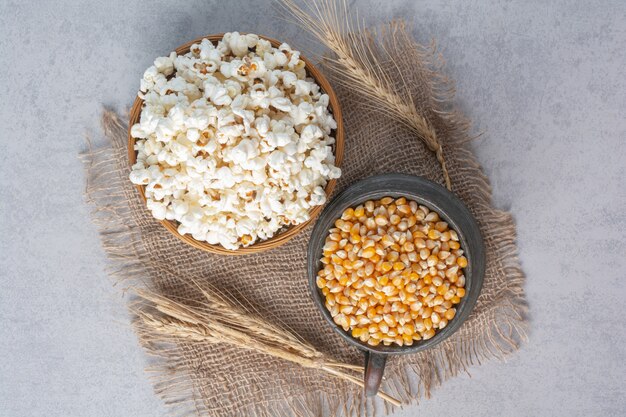 Dzbanek wypełniony kukurydzą i miska wypełniona popcornem obok łodyg pszenicy na kawałku tkaniny na marmurze.