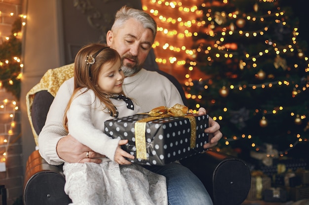 Dziadek siedzi z wnuczką. Świętowanie Bożego Narodzenia w przytulnym domu. Mężczyzna w białym swetrze z dzianiny.