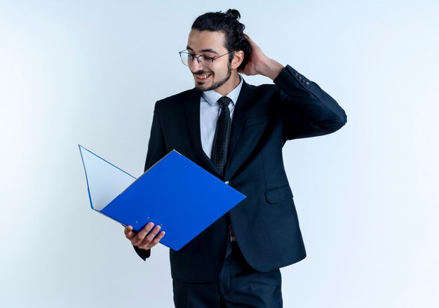 Działalności człowieka w czarnym garniturze i okularach, trzymając folder patrząc zdezorientowany drapiąc się po głowie stojąc na białej ścianie
