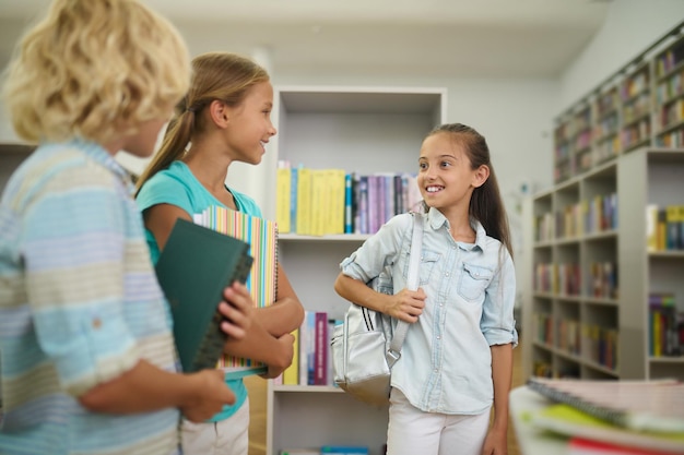 Dzieci, biblioteka. Dwie uśmiechnięte długowłose dziewczyny z plecakiem i zeszytami oraz blond chłopiec z książkami stoją rozmawiając w jasnym pokoju w bibliotece