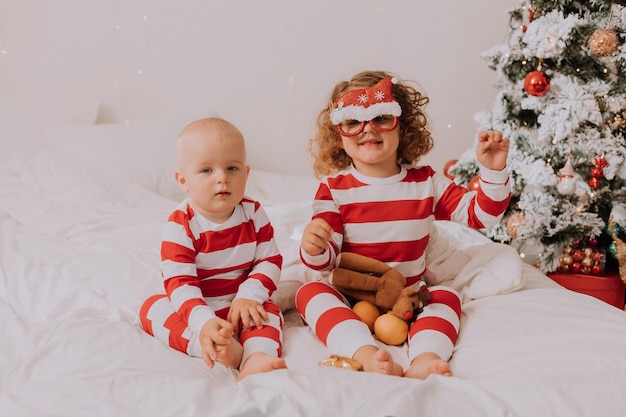 Dzieci w biało-czerwonych piżamach przymierzają śmieszne okulary z mikołajem siedzącym w łóżku. styl życia. brat i siostra świętują boże narodzenie. chłopiec i dziewczynka bawią się w domu. zdjęcie wysokiej jakości
