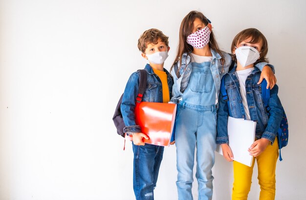 Dzieci w maskach trzymające szkolne rzeczy i stojące pod ścianą – COVID-19