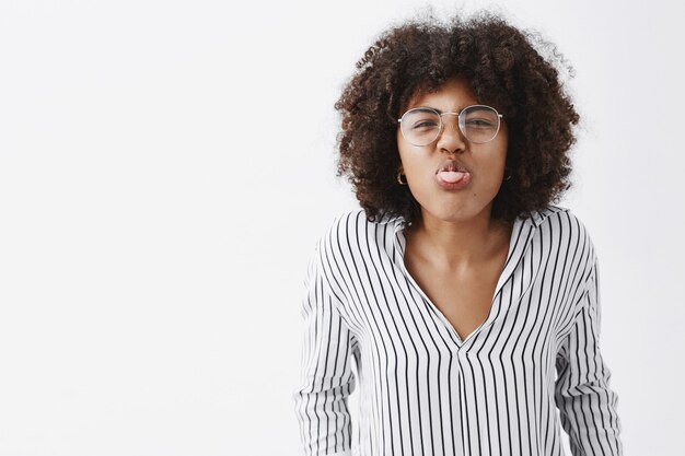 dziecinna i figlarna zabawna afroamerykańska dorosła kobieta z fryzurą afro w biurowej bluzce w paski i okularach pochylająca się w kierunku i pokazująca język radośnie marszczący nos