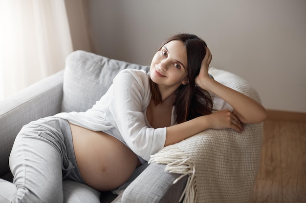 Dziecko chce, abyś dotykał brzucha Portret szczęśliwej, zrelaksowanej ciężarnej europejskiej kobiety w wygodnym, wygodnym stroju, siedzącej w fotelu opierając się na dłoni i patrzącej na aparat z uroczym uśmiechem