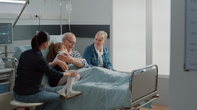 Bezpłatne zdjęcie dziecko przytula chorego dziadka w łóżku na oddziale szpitalnym podczas wizyty, pocieszając starzejącego się pacjenta z chorobą. dziecko i matka odwiedzają starszego mężczyznę z woreczkiem do kroplówki i nosową rurką tlenową. odwiedzający w klinice