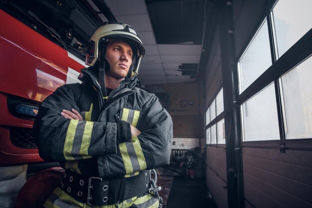 Dzielny strażak w mundurze ochronnym stojący obok wozu strażackiego w garażu straży pożarnej ze skrzyżowanymi rękami i patrzącym w bok
