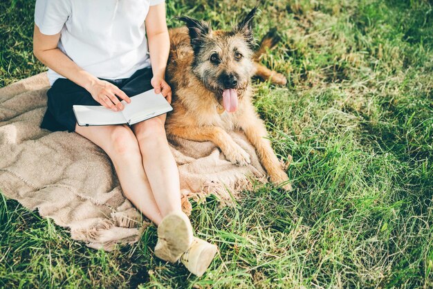 Dziewczyna czyta książkę, siedząc na kocu w pobliżu dużego starego psa na trawniku zielonej trawie w słonecznym parku publicznym w miły dzień.