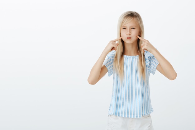 Dziewczyna dąsająca się, wyglądająca jak balon. Portret ślicznej dziecinnej młodej blond córki w niebieskiej stylowej bluzce, dąsającej się i wskazującej na policzki, nieposłusznej i znudzonej, stojącej nad szarą ścianą
