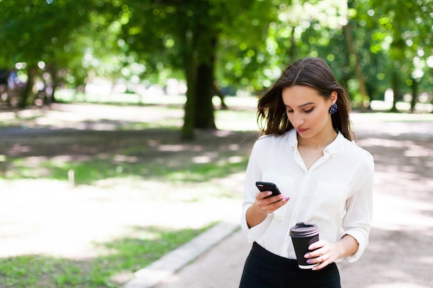 Dziewczyna idzie z telefonu w ręce i filiżankę kawy w parku