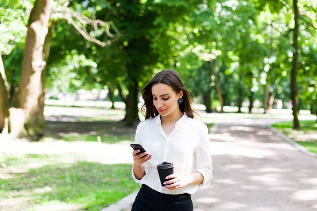 Dziewczyna idzie z telefonu w ręce i filiżankę kawy w parku