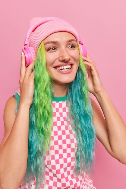 Bezpłatne zdjęcie dziewczyna lubi ścieżkę dźwiękową w bezprzewodowych słuchawkach dreszcze w pomieszczeniu słucha muzyki z głośnym dźwiękiem ma kolorowe farbowane włosy nosi kapelusz i kraciastą sukienkę na różowo