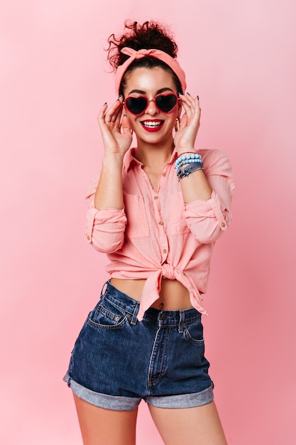 Dziewczyna pin-up w opasce zakłada okulary przeciwsłoneczne w kształcie serca. Portret kobiety w Dżinsowe szorty i koszulę, pozowanie na różowej przestrzeni.