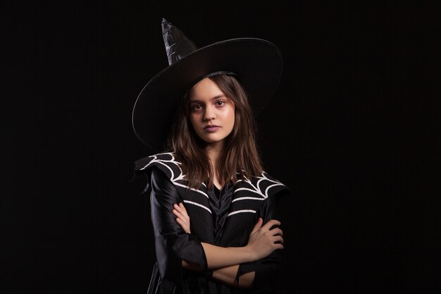Dziewczyna robi ciemne zaklęcia ze skrzyżowanymi rękami i poważnym wyrazem twarzy na karnawał w halloween. Młoda czarownica robi ciemne czary.