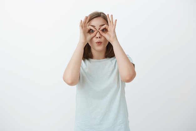 Dziewczyna robi śmieszne miny marnuje czas. Portret figlarnej i radosnej niedojrzałej uroczej kobiety w t-shircie, która krąży wokół oczu rękami, jakby patrzyła przez gogle składane usta, wygłupiając się