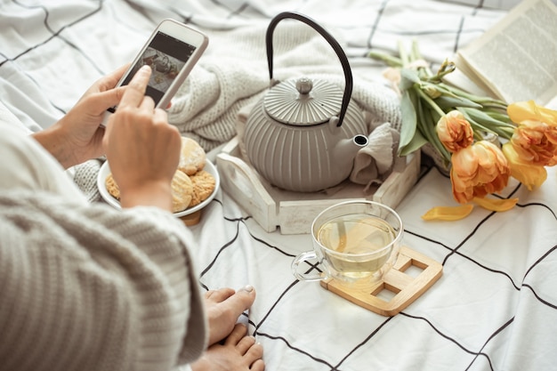 Dziewczyna robi zdjęcia telefonem wiosennej kompozycji z herbatą, ciasteczkami i tulipanami w łóżku