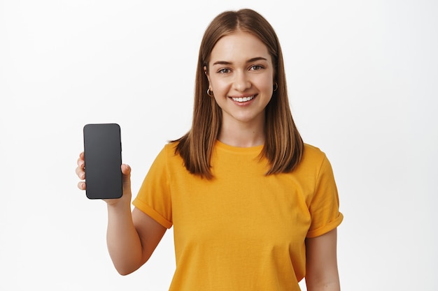 Dziewczyna trzyma smartfon i uśmiecha się, pokazując aplikację interfejsu, pusty ekran telefonu komórkowego, stojąc w żółtej koszulce nad białą ścianą.