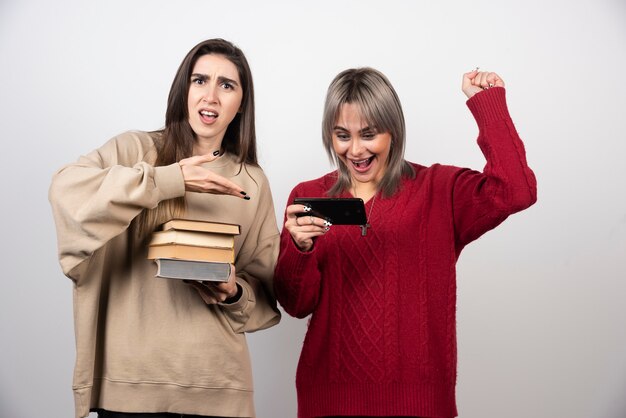 Dziewczyna w beżowym swetrze trzyma książki, podczas gdy inna dziewczyna patrzy na telefon.