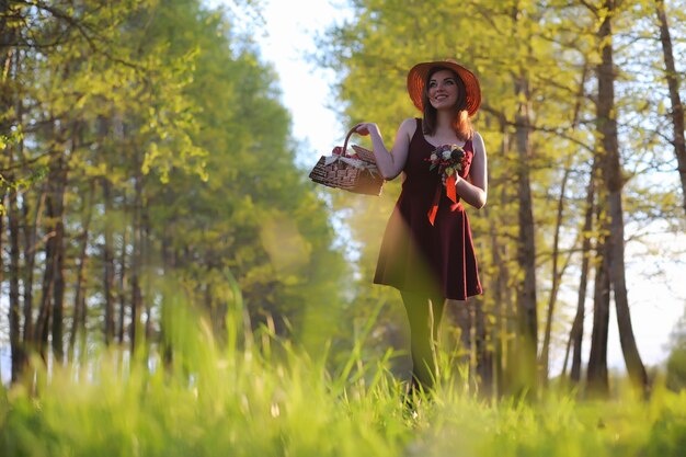 Dziewczyna w kapeluszu na spacerze w parku. dziewczyna z koszykiem spaceruje wiosną. dziewczyna idzie drogą o zachodzie słońca.