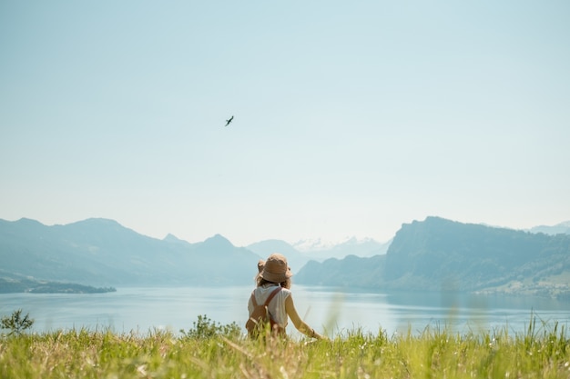 Dziewczyna w kapeluszu siedzi na zielonym trawniku w pobliżu jeziora