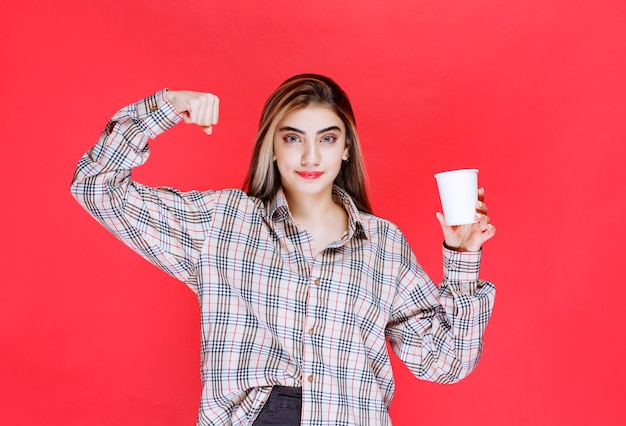 Dziewczyna w kraciastej koszuli trzymająca białą, jednorazową filiżankę kawy i pokazująca swoją moc