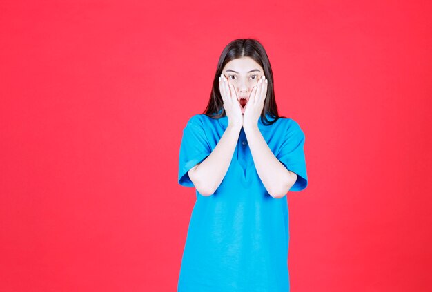 Dziewczyna w niebieskiej koszuli stoi na czerwonej ścianie i wygląda na przestraszoną i przerażoną