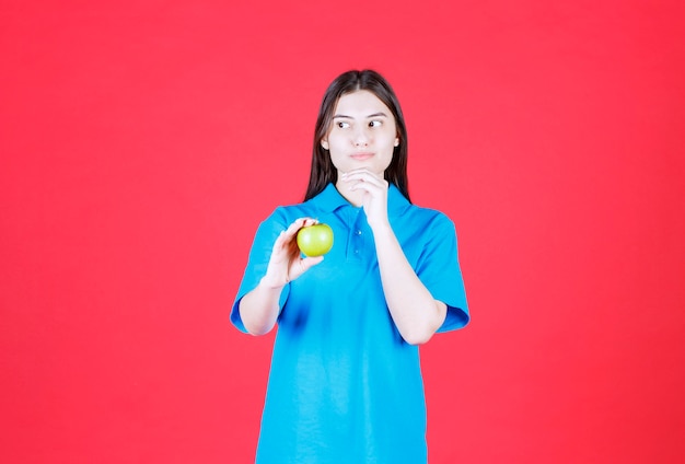 Dziewczyna w niebieskiej koszuli trzymająca zielone jabłko i myśląca i wahająca się
