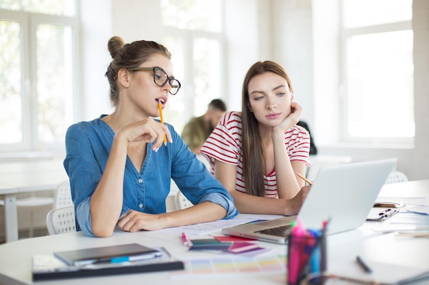 Dziewczyna w okularach z ołówkiem i zamyślona dziewczyna w koszulce w paski, opierając się na dłoni podczas zamyślonej wspólnej pracy z laptopem Młode kobiety spędzające czas w nowoczesnym biurze