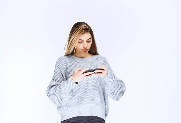 Bezpłatne zdjęcie dziewczyna w szarej bluzie trzymająca czarny smartfon i pisząca wiadomość lub komentarz.
