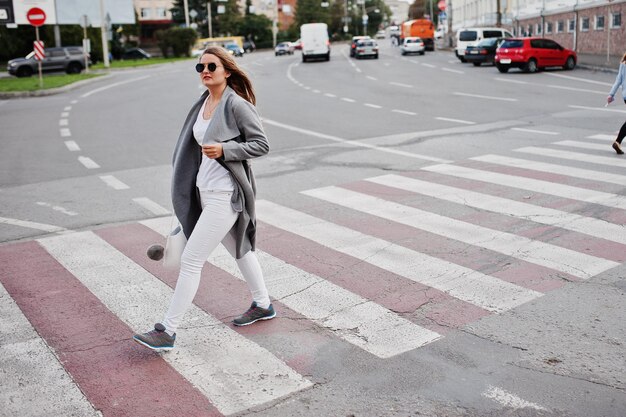 Dziewczyna w szarym płaszczu z okularami przeciwsłonecznymi i torebką spacerująca po przejściu dla pieszych