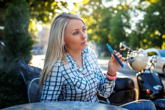 Dziewczyna z długimi blond włosami w tshirt, siedząc przy stole na zewnątrz i paląc elektroniczny papieros.