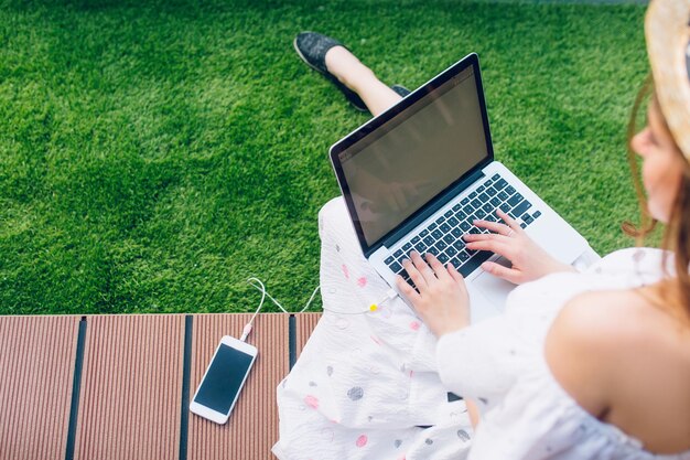 Dziewczyna z długimi włosami w kapeluszu siedzi na podłodze z desek. Nosi białą sukienkę z odkrytymi ramionami. Pisze na laptopie na kolanach. Widok z góry, skupienie się na laptopie