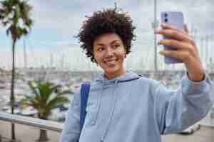 Bezpłatne zdjęcie dziewczyna z kręconymi włosami ubrana w luźną bluzę z kapturem robi selfie portret na smartfonie spaceruje po porcie cieszy się dobrym dniem