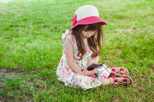 Dziewczynka siedzi na trawie, patrząc na telefon komórkowy ze słuchawkami