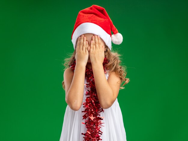 Dziewczynka ubrana w świąteczny kapelusz z girlandą na szyi zakrytej twarzy rękami odizolowanymi na zielonej ścianie