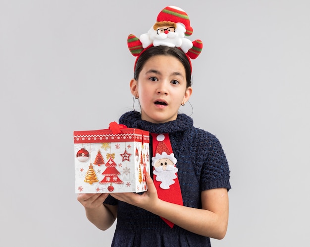 Dziewczynka w dzianinowej sukience ubrana w czerwony krawat z zabawną świąteczną obwódką na głowie trzyma prezent na Boże Narodzenie, patrząc zaskoczony