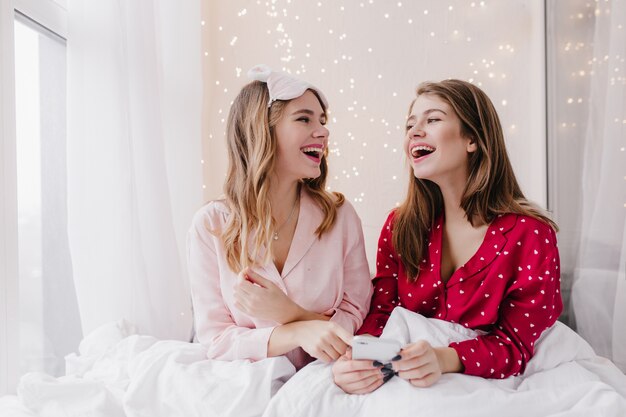 Dziewczyny w piżamach patrzą na siebie i uśmiechają się. Kryty zdjęcie roześmianych kaukaskich pań siedzących w łóżku ze smartfonem.