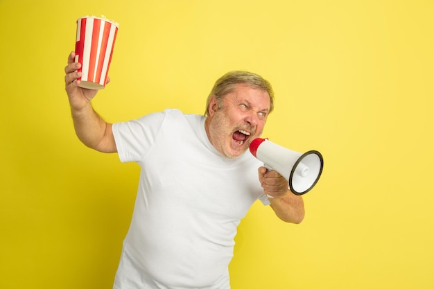 Dzwoni z spokojem ust, trzyma popcorn. Portret mężczyzny kaukaski na żółtym tle studio. Piękny model męski w białej koszuli.