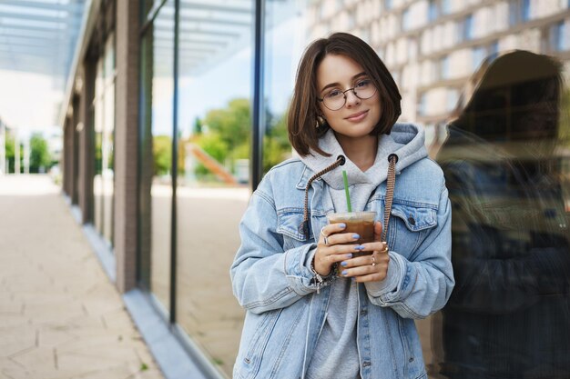 Edukacja kobiety i koncepcja stylu życia Portret delikatnej hipsterki spacerującej samotnie po mieście, patrząc romantycznie i szczęśliwie, uśmiechnięta kamera opiera się na budowaniu i piciu kawy z lodem latte