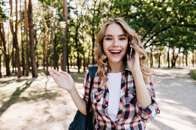 Ekstatyczna biała dziewczyna rozmawia przez telefon podczas odpoczynku w lesie. Zewnątrz zdjęcie radosnej kobiety z falowanymi włosami spacerującej po parku.