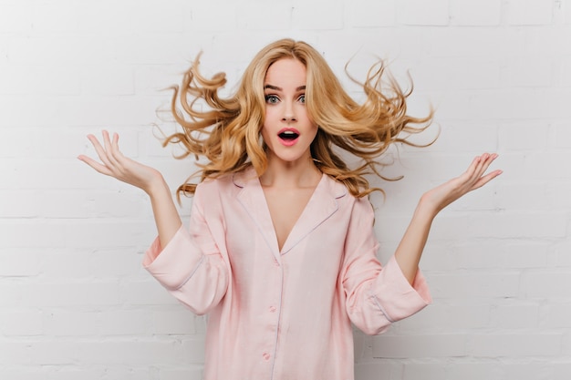 Ekstatyczna niebieskooka kobieta z długimi blond włosami pozuje przed białą ceglaną ścianą. Kryty strzał zdziwionej dziewczyny w pięknej różowej piżamie.