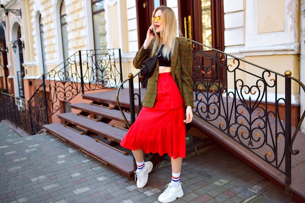 Elegancka modna blondynka pozuje na ulicy w pobliżu pięknego starego budynku, mówiąc przez telefon, ubrana w modny modny strój hipster i okulary przeciwsłoneczne, styl wiosna jesień.