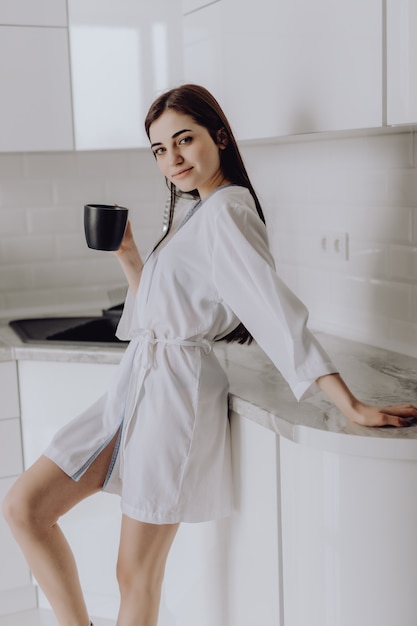 Elegancka uśmiechnięta kobieta w białym szlafroku stoi przed kuchnią, picie porannej kawy