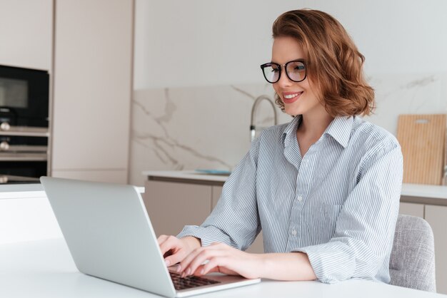 Elegancka uśmiechnięta kobieta w szkłach i pasiastej koszula używać laptop podczas gdy będący usytuowanym przy stołem w kuchni