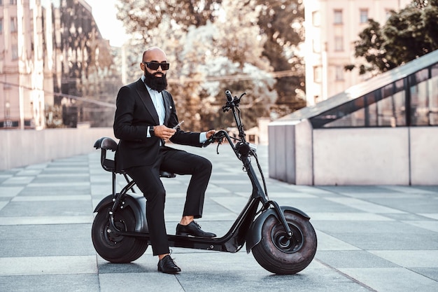 Elegancki łysy biznesmen w okularach przeciwsłonecznych pali waporyzator siedząc na swoim elektrycznym rowerze.