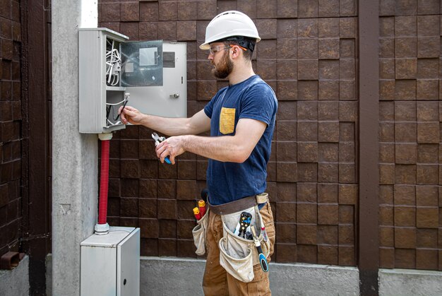 Elektryk konstruktor przy pracy, bada połączenie kablowe w linii elektrycznej w kadłubie przemysłowej rozdzielnicy. Profesjonalista w kombinezonie z narzędziem elektryka.