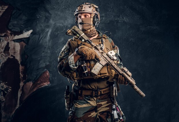 Elitarna jednostka, żołnierz sił specjalnych w mundurze kamuflażowym pozujący z karabinem szturmowym. Zdjęcie studyjne na tle ciemnej, teksturowanej ściany