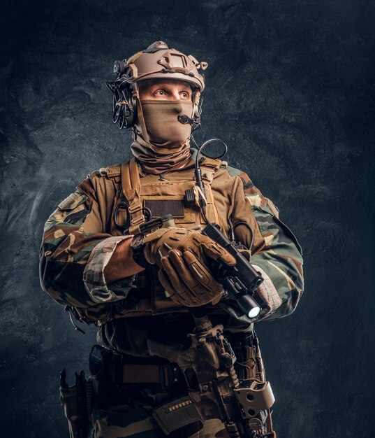Elitarna jednostka, żołnierz sił specjalnych w mundurze kamuflażowym trzymający broń z latarką i patrzący w bok. Zdjęcie studyjne na tle ciemnej, teksturowanej ściany