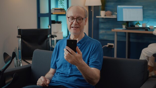 Emerytowany mężczyzna trzymający smartfona na czacie podczas rozmowy wideo z rodziną, przy użyciu telekonferencji online do komunikacji na odległość. Emeryt rozmawiający z ludźmi na spotkaniu wideokonferencyjnym.